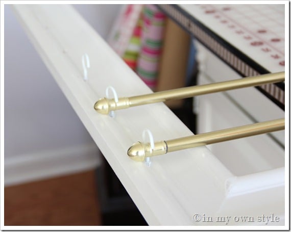 White cup hooks in white frame and brass curtain rods in cup hooks. How to organize gift wrap