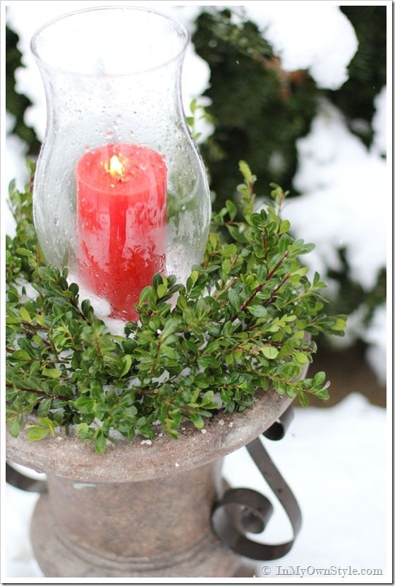 Red candle in glass hurricane on outdoor planter urn.