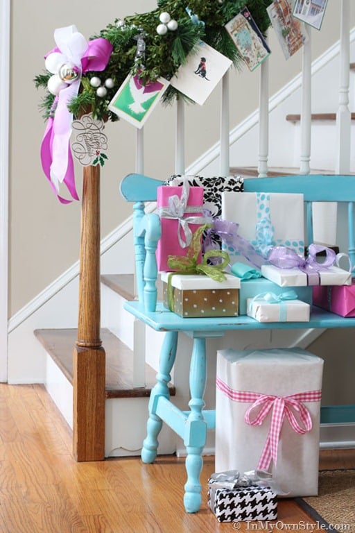 Christmas garland with cards tied on draped on a staircase railing. Turquoise bench with wrapped Christmas presents in hallway.