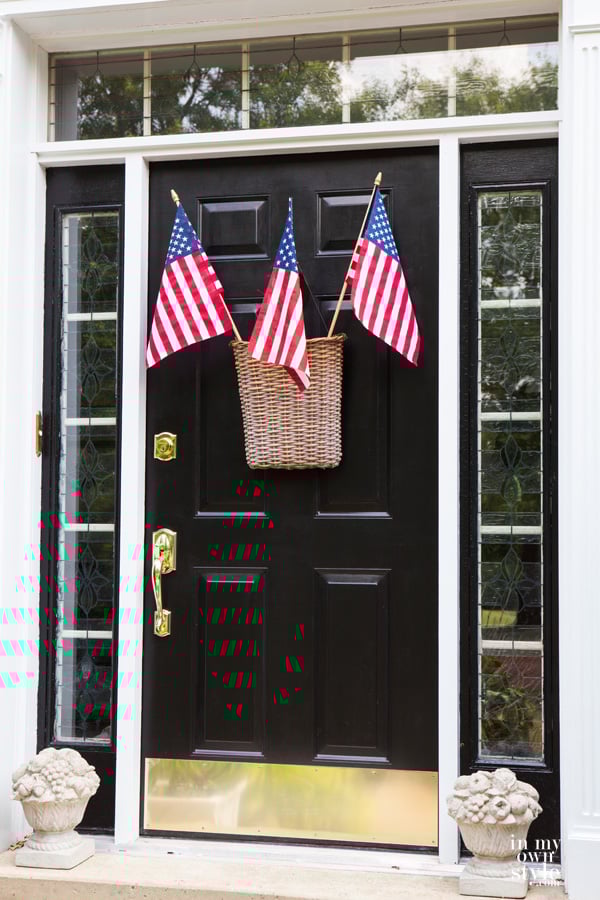 Door-Decoration-Ideas-for-the-Fouth-of-July-and-Patriot-holidays - 3 American flags sticking out of a flat backed rattan hanging basket on the front door of a house.