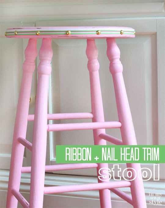 Pink painted stool with ribbon and nail head trim around the edge of the seat.