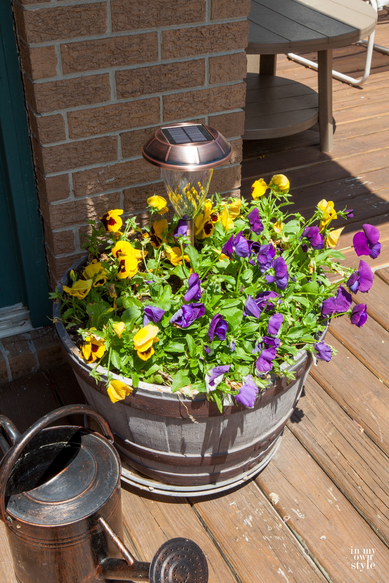 Copper solar light in pansy filled planter. Watering can next to it. Another way to add outdoor deck lighting in an easy way.