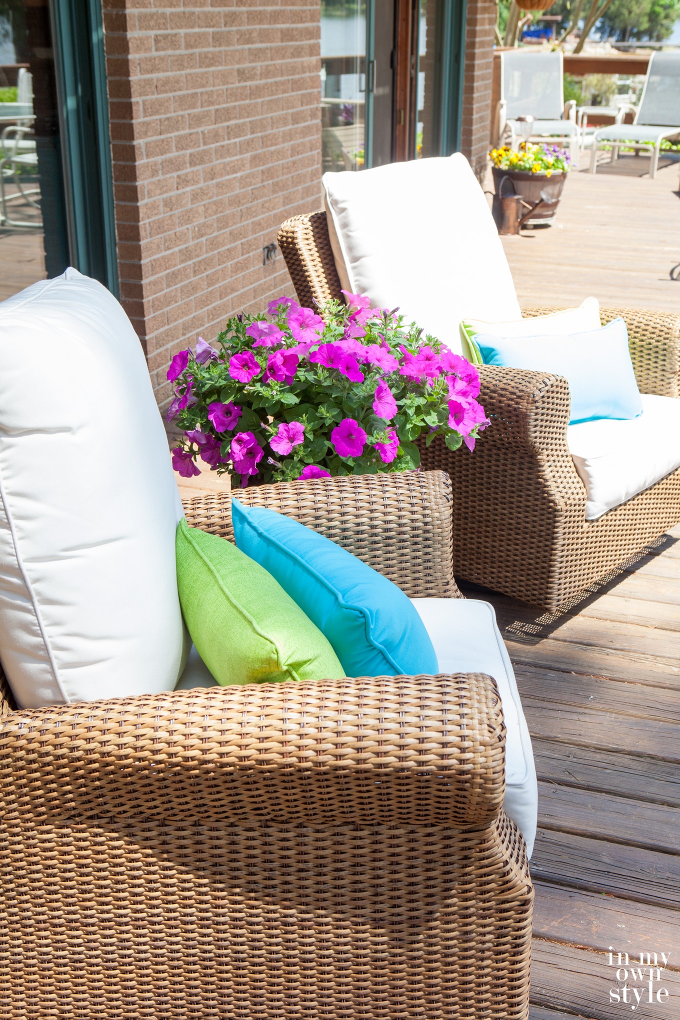 Two rattan chairs with white cushions on deck. Pink Petunias on table.