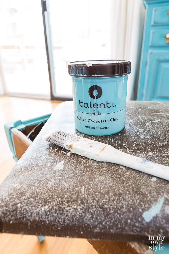 An plastic gelato container being used to hold turquoise chalk paint. It is placed on the unfinished stool with a paint brush.