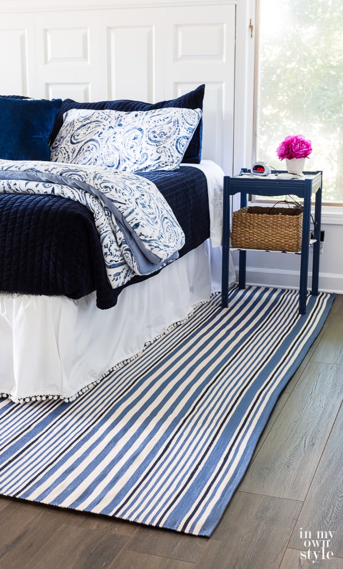 Blue and white bedroom with blue and shite striped area rug over dark hardwood floor.