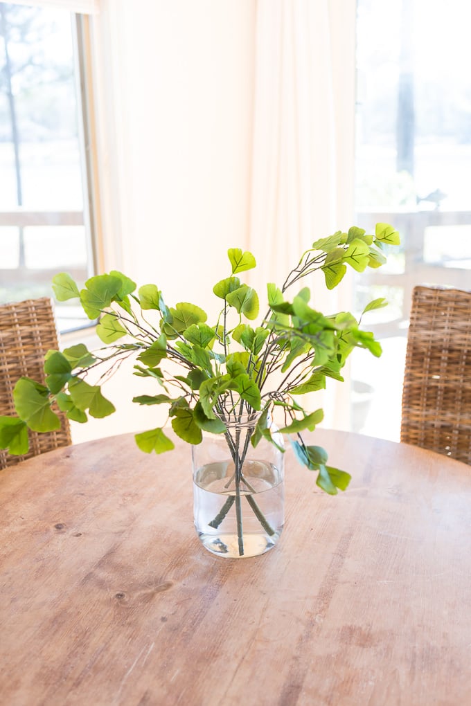 Fake Gingko house plant in large clear glass vase filled with water on top of a round wood table.