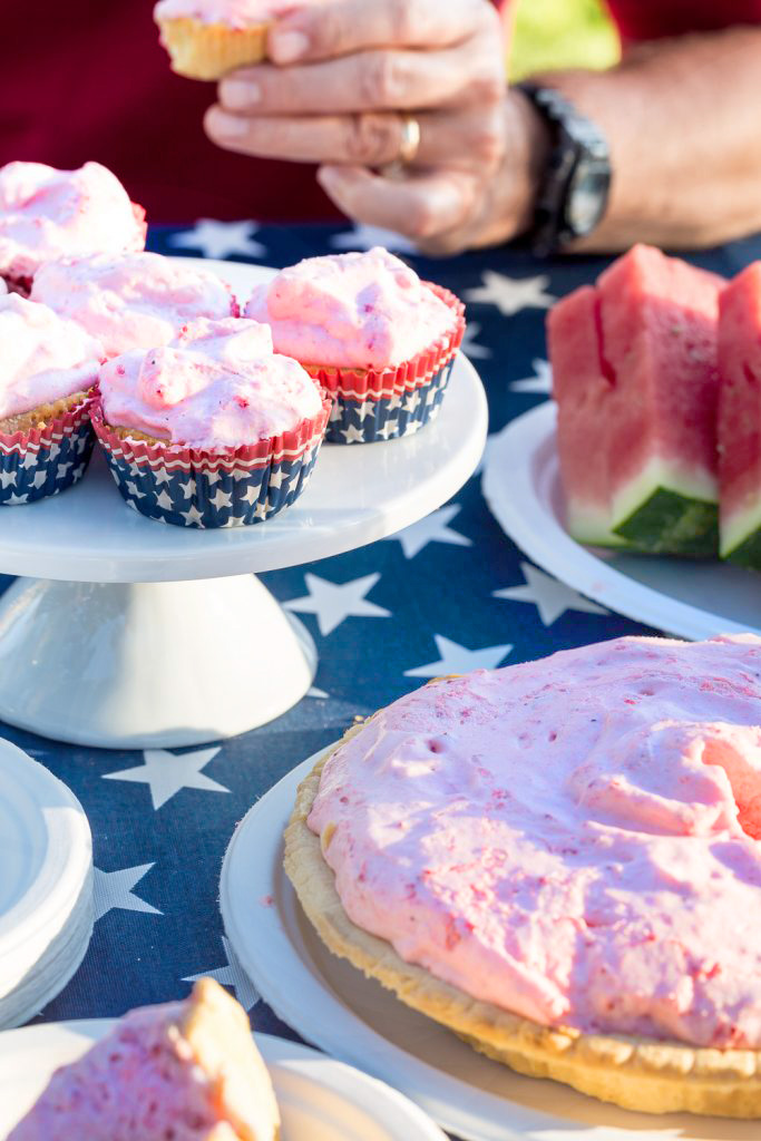 Frozen strawberry cream pie set on a patriotic table cloth covered table set up for an outdoor picnic. Man's hand holding a pie cupcake.