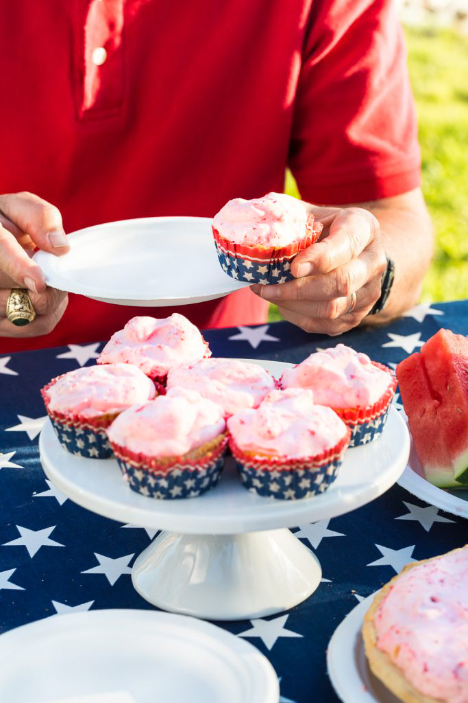 Frozen strawberry cupcakes in patriotic paper cupcake liners on white cake stand. Man's hand lifting one to place on plate.