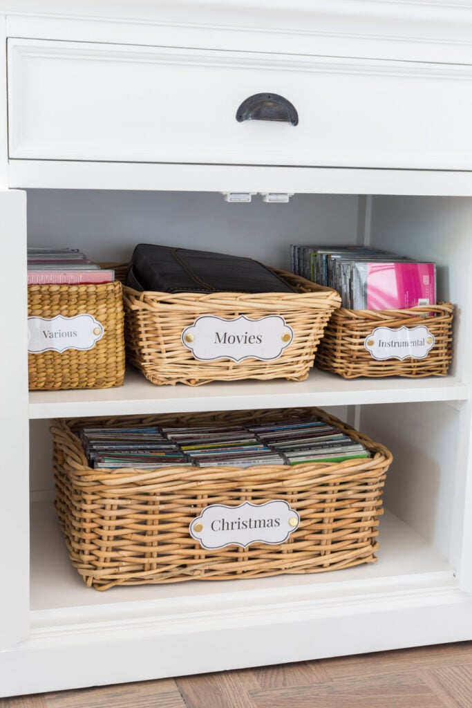 Storage baskets with laminated DIY labels arranged on the shelves inside a storage cabinet.