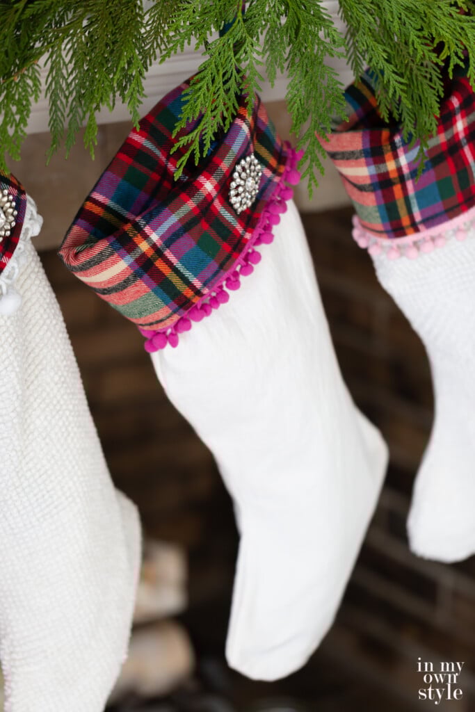 Pink, green and red plaid trimmed Christmas stocking with rhinestone pin hung on a mantel