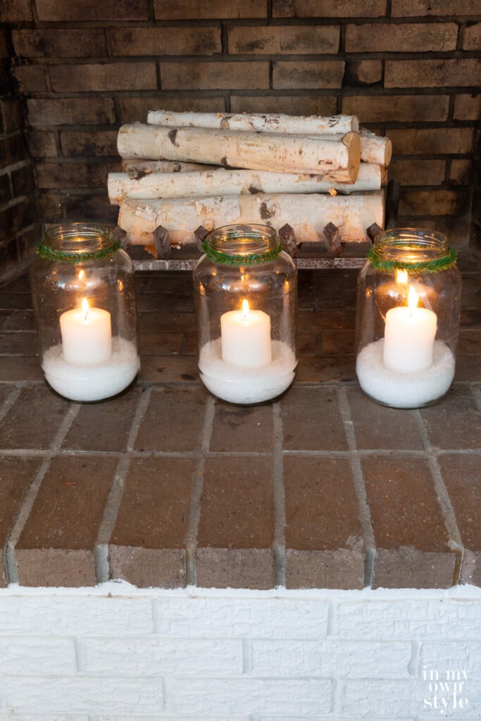 3 large glass jars filled with fake snow and white candles in a fireplace.