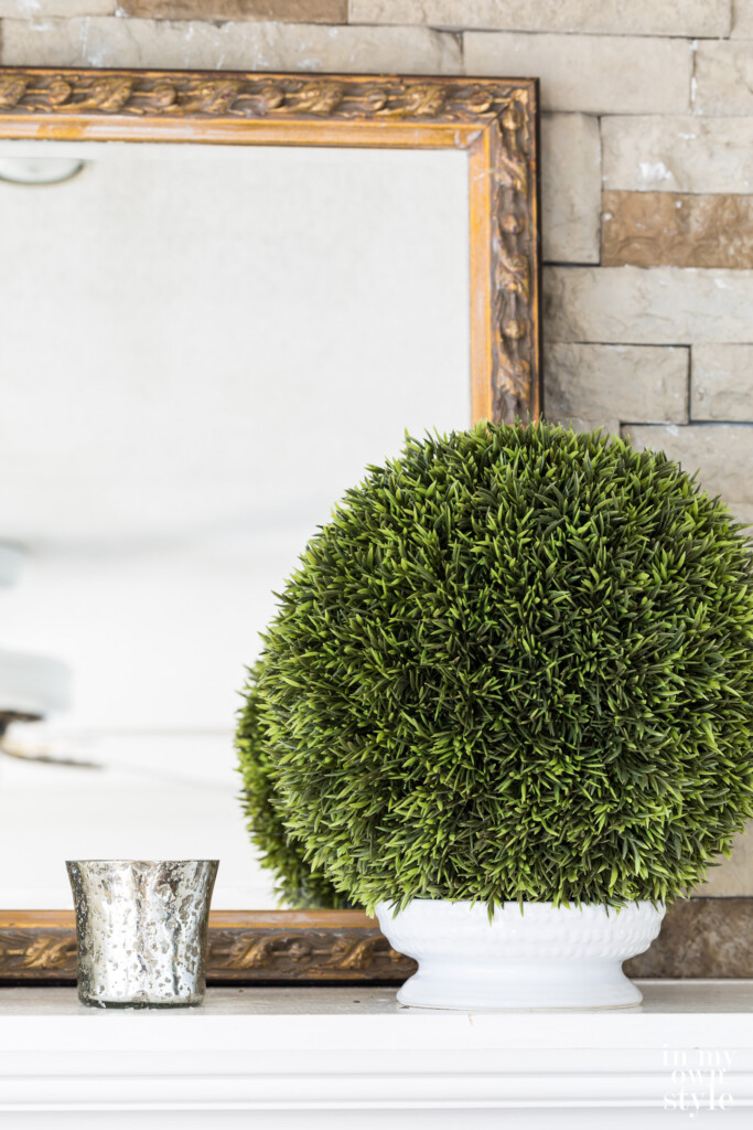 Mantel with a gold framed mirror, topiary ball and silver votive candle holder