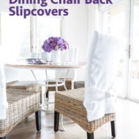 Casual dining area in a home with table and chairs. Chairs are wicker and have white chair back slipcovers on them. A white pitcher on the table has purple hydrangeas in it.