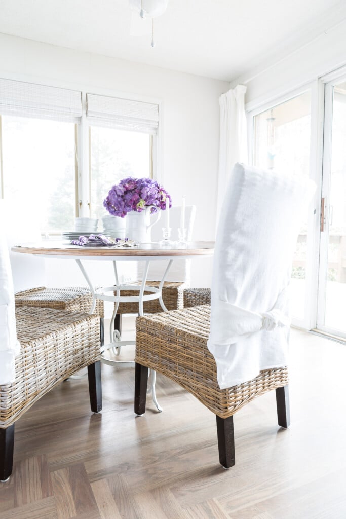 Casual and simple spring decorating for a dining room. Rattan chairs with white slipcovers on the backs. Round wood table with white wrought iron base.