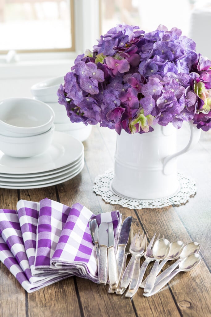 Spring home decor. Decorating for spring using White dinnerware and white pitcher filled with purple hydrangeas on a wood table. Stack of white plates and bowls along the side of the pitcher.