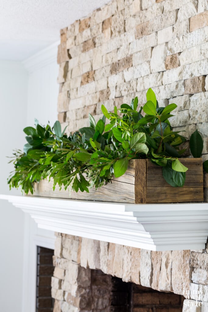 White mantel on stone fireplace with wood planter box filled with greenery.