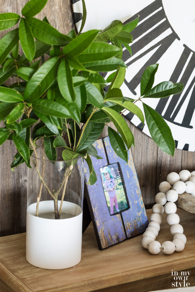 Summer decorating ideas for a sofa table. Green clippings from shrubs in glass and white vase next to purple photo frame and wood beads on a table.