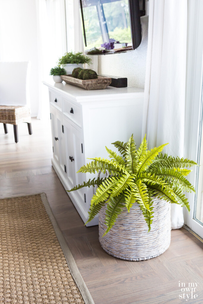 Large white washed basket with a faux fern in it in a living room.
