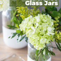 Two pickle jars on table that were dipped in white paint to make modern looking vases. Text overlay says Creative ways to reuse glass jars.