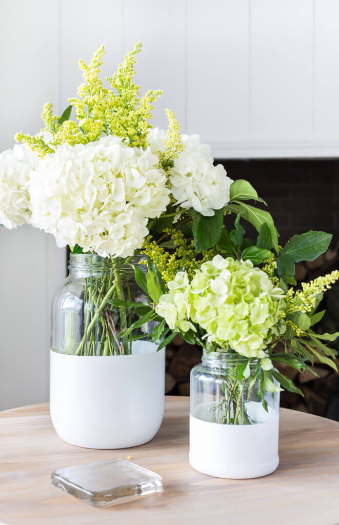 Two paint dipped glass pickle jars turned into chic and modern vases. They are placed on a wood side table.