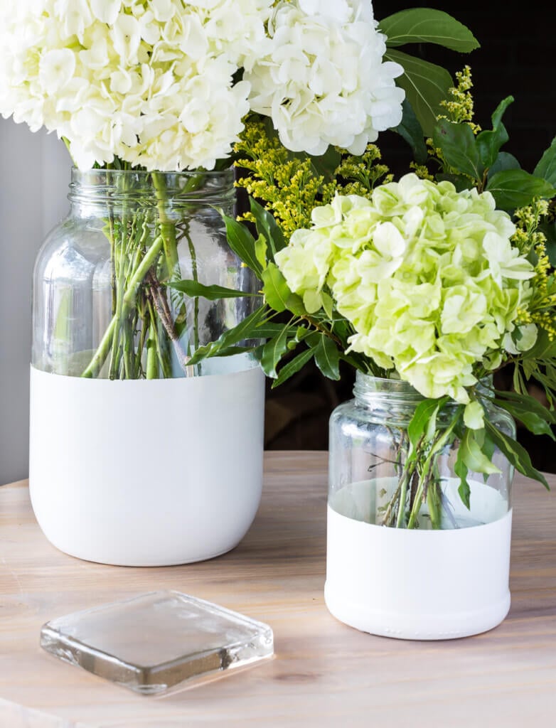 Two sizes of glass pickle jars transformed into modern white dipped glass vases. They are displayed on a side table.