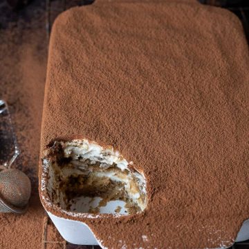 A white dish of vegan tiramisu with a spoonful removed on a wire rack on a dark background dusted with cocoa powder.