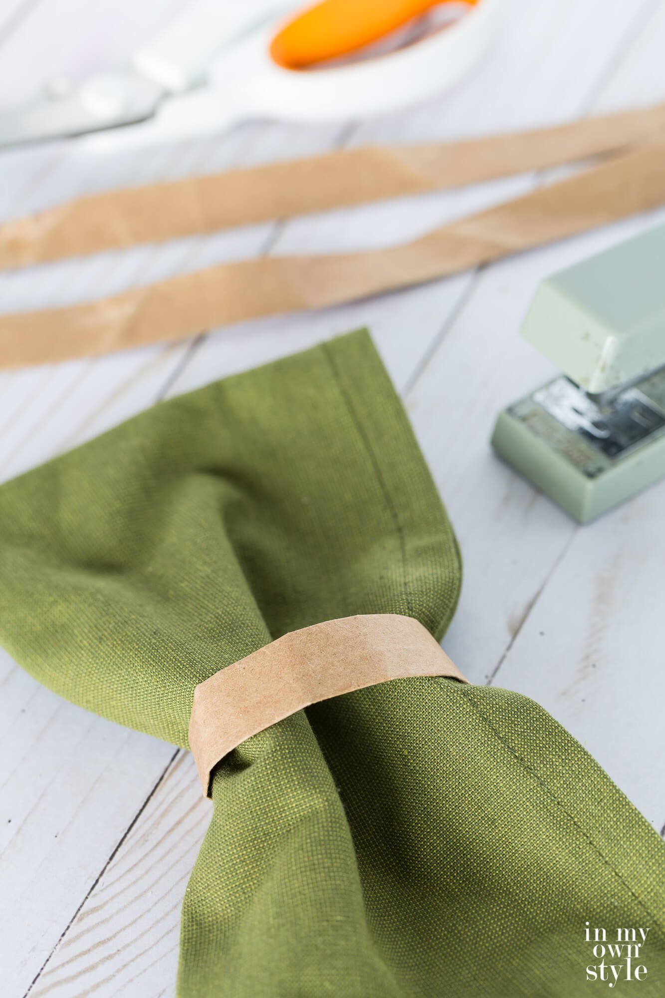 Brown paper bag handle napkin ring on dark sage green napkin. Stapler and scissors in the background.