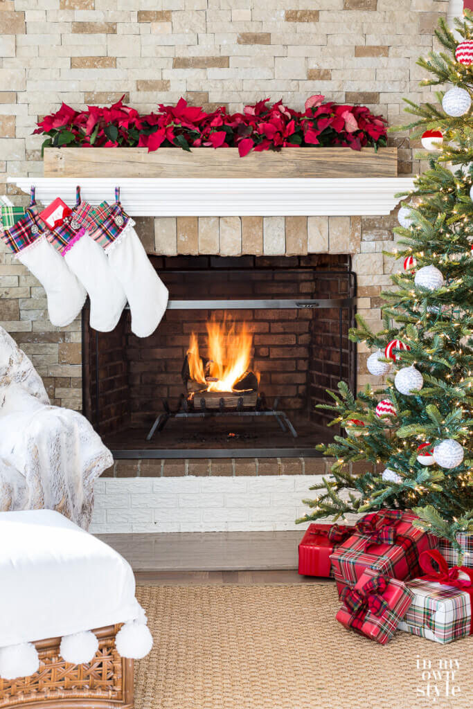Stone fireplace with white stockings hung on the left. Christmas tree on right. Fire in fireplace.