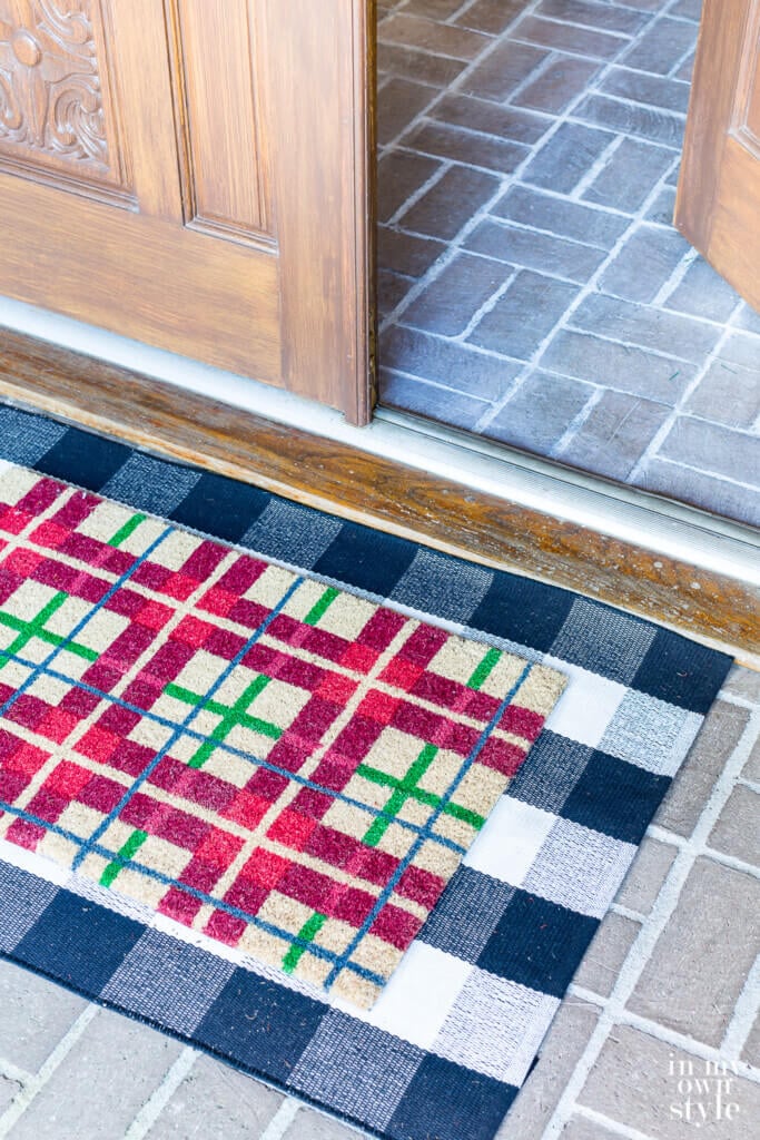 red and green plaid doormat on top of a black and white check mat by front door of holiday decorated home