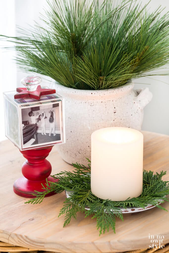 A red pedestal photo cube, a candle on greenery and a planter with pine on a table.