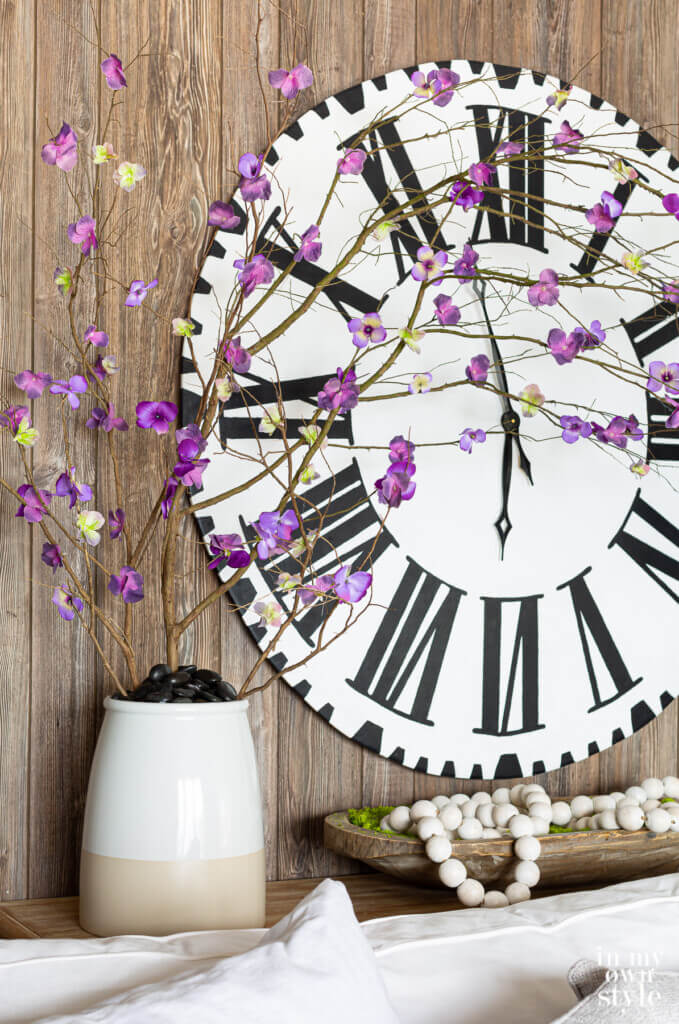 Large Roman numeral wall clock on wall with a ceramic planter filled with faux forced branches with purple flowers.