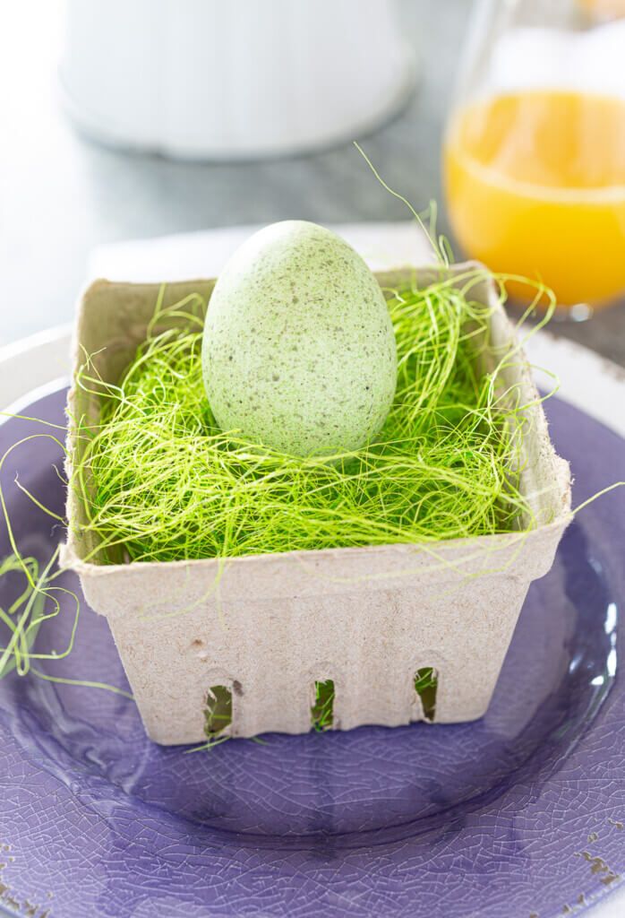 Cardboard berry crate filled with green Easter grass and a pale green speckled egg. Sitting on the center of a purple dinner plate.