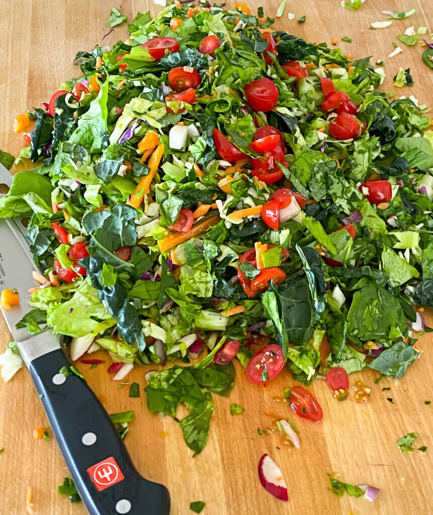 Cut up lettuce . leafy greens and vegetables on a wood cutting board. Chef's knife on the side.