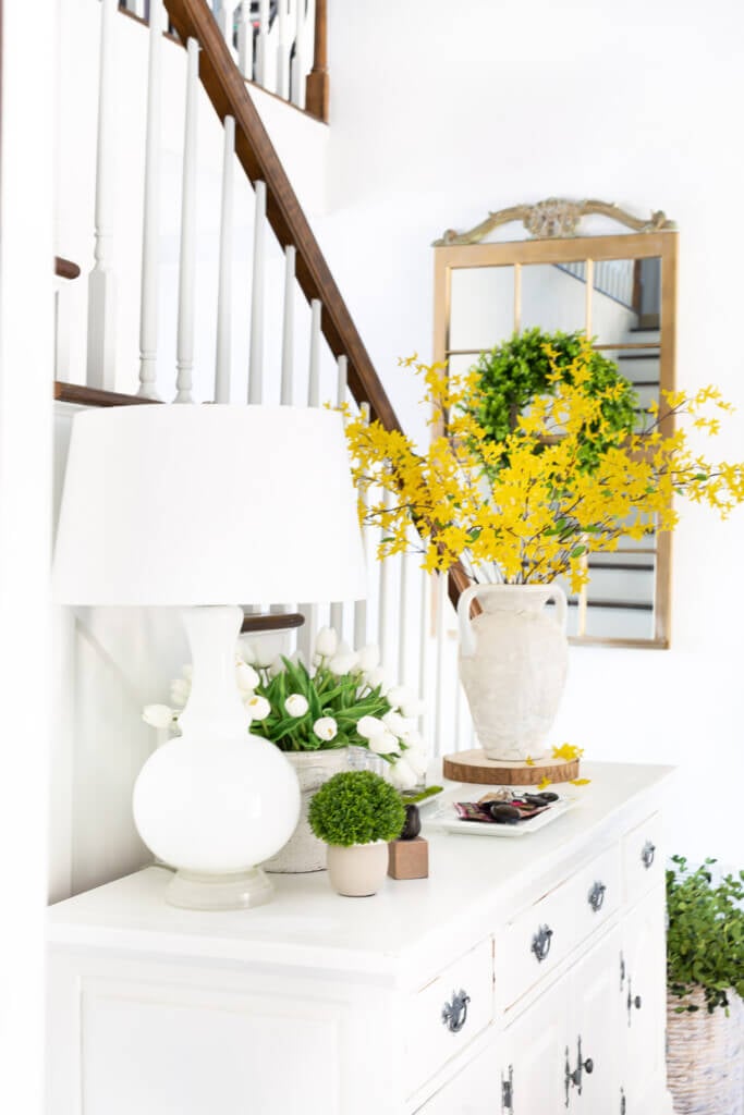 Forsythia and white tulips on a long white dresser in entryway foyer decorated for spring.