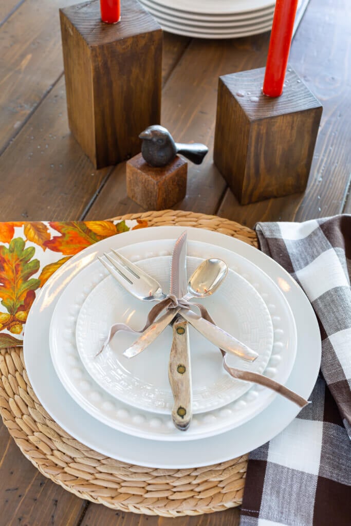 Table place setting of a white dinner plate layered with a fork, knife and spoon tied with brown ribbon.