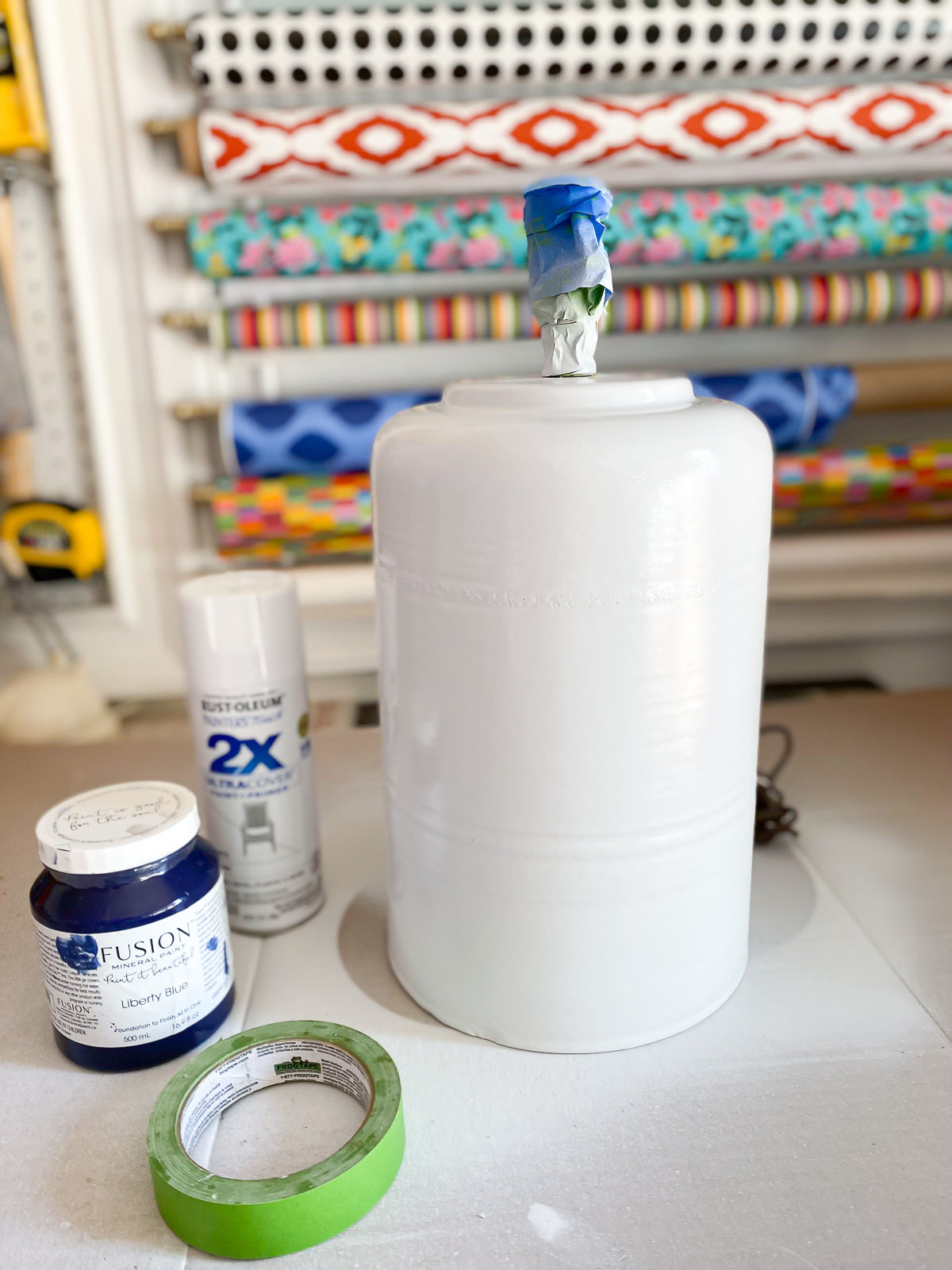 White spray painted lamp base on work table with paints used next to it.