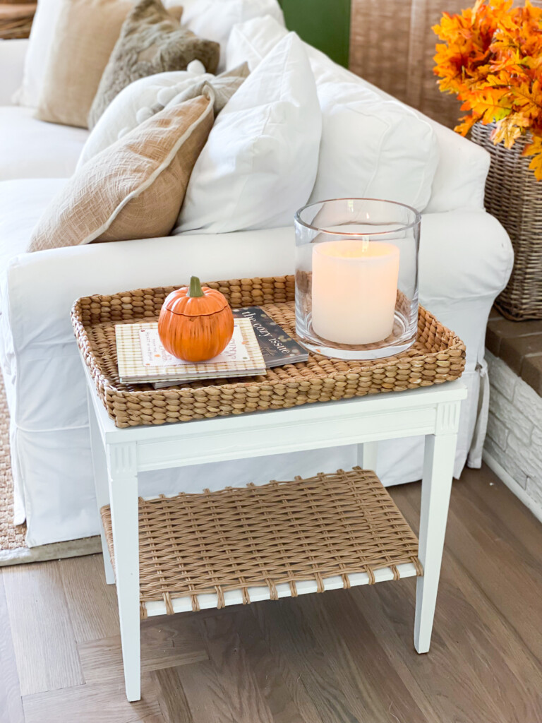 Two tier white side table with woven tray a small pumpkin and candle in glass hurricane