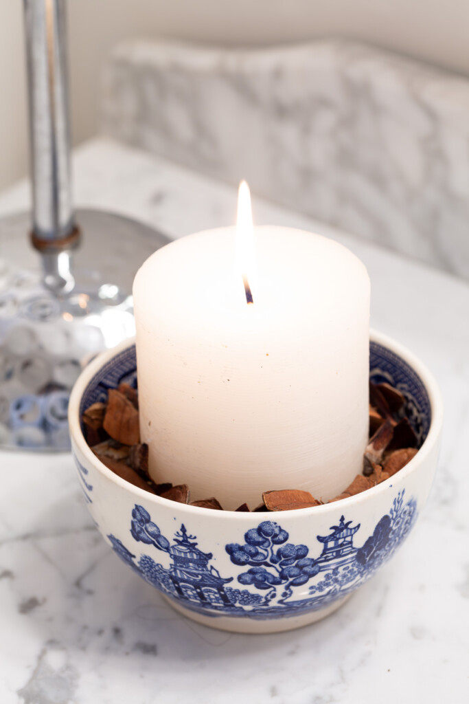 White candle in a bed of pine cone needles in.a blue and white bowl.