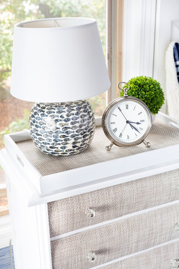 A night table with a table lamp on a round wood riser, silver alarm clock and a small plant.