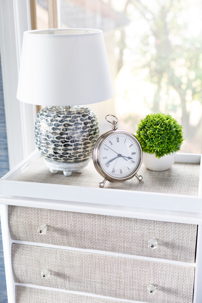 A night table with a table lamp on a round wood riser, silver alarm clock and a small plant.