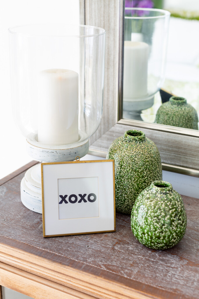 XOXO in a frame with green and white vases and white candle holder on a table top.