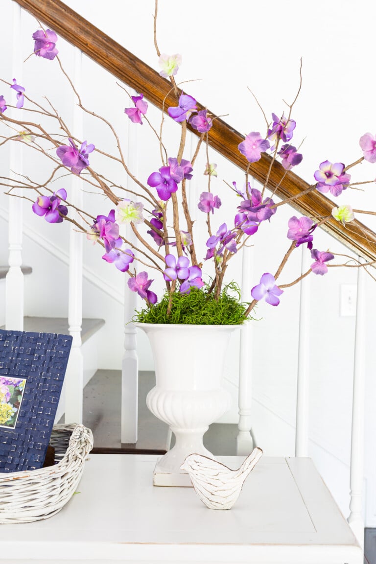 faux purple flowering branches in a white ceramic urn on a sideboard.
