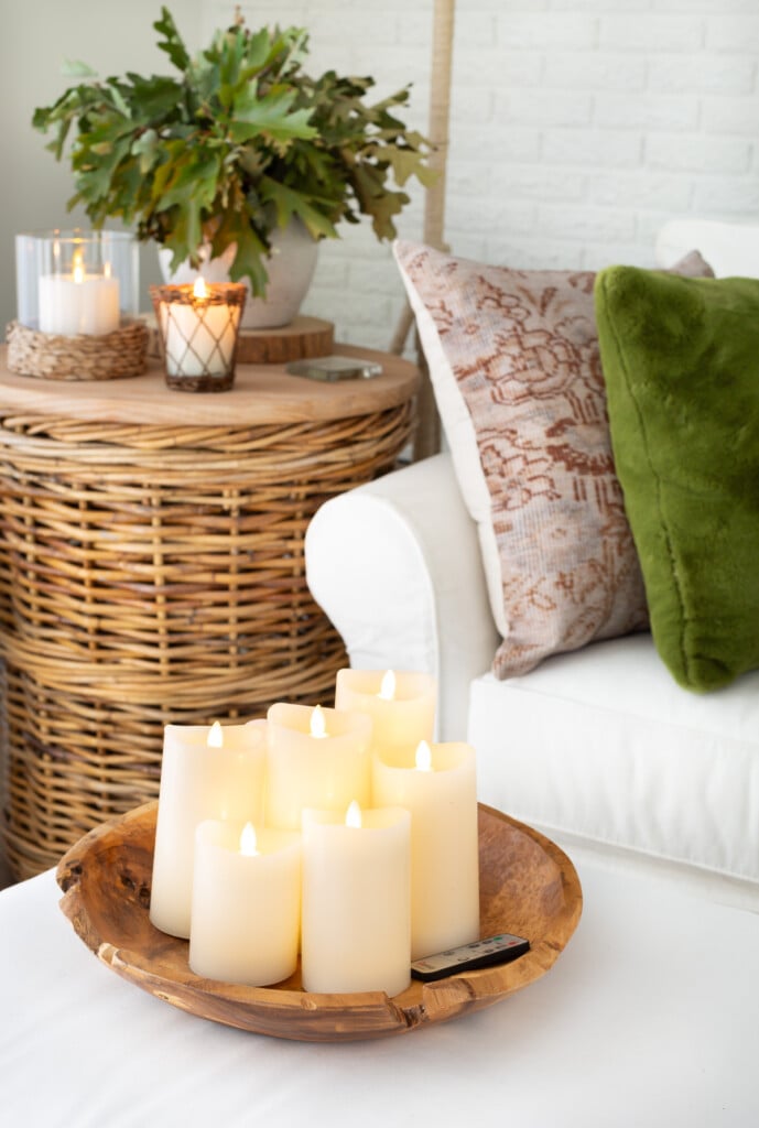 Large wood bowl on ottoman filled with battery operated off-white pillar candles in a living room.