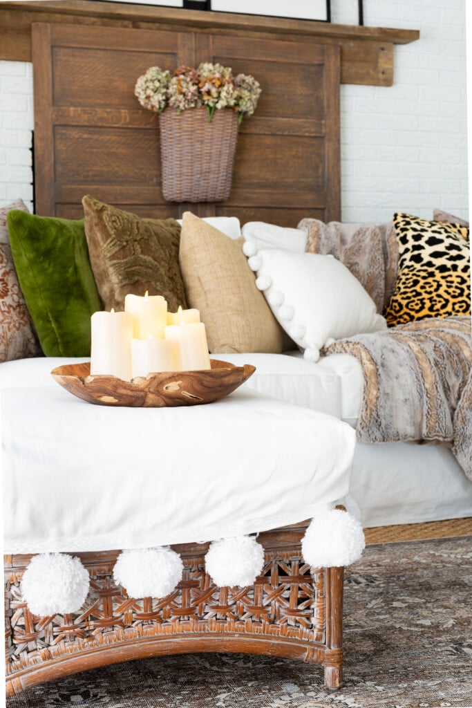 Large wood bowl on ottoman filled with battery operated off-white pillar candles in a living room.