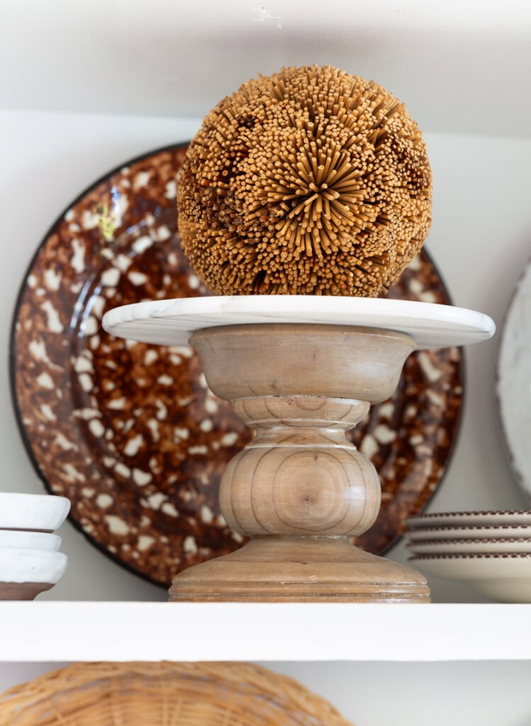 A small wood pedestal with a decorative fall colored orb on am open shelf in a kitchen.