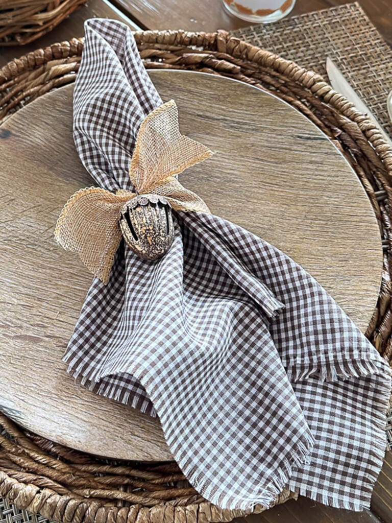 brown and white gingham napkin tied with burlap ribbon and a metal acorn jingle bell on wood look dinner plate.