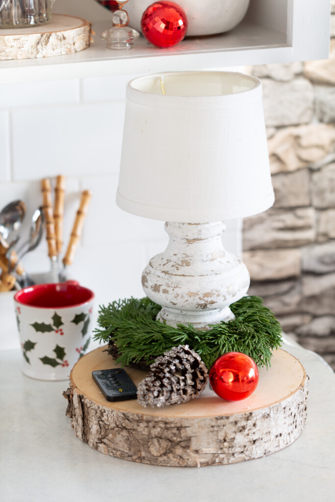 small lamp on kitchen counter with evergreen wreath, a glittered pinecone, and a red ball ornament surrounding it.