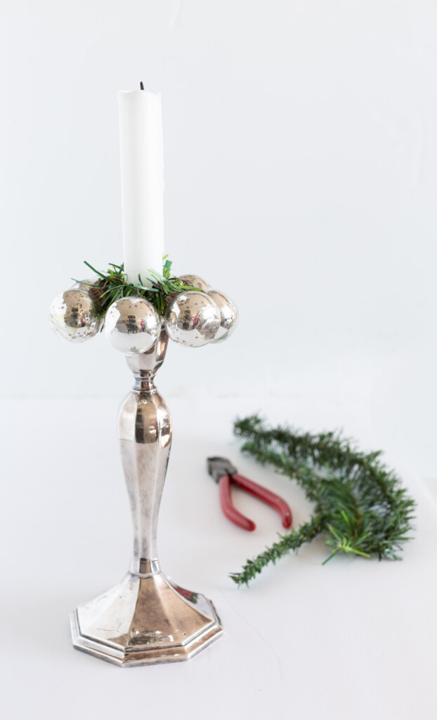 Tall silver candlestick holder and white candle. The Christmas candle ring is made using small silver ornaments and a garland tie.