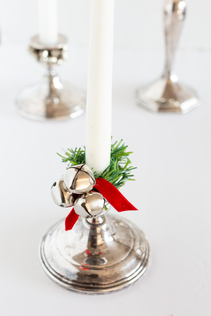 Silver candle holder with a white candle with a faux greenery garland tie, jingle bell and red ribbon made into a taper candle ring.