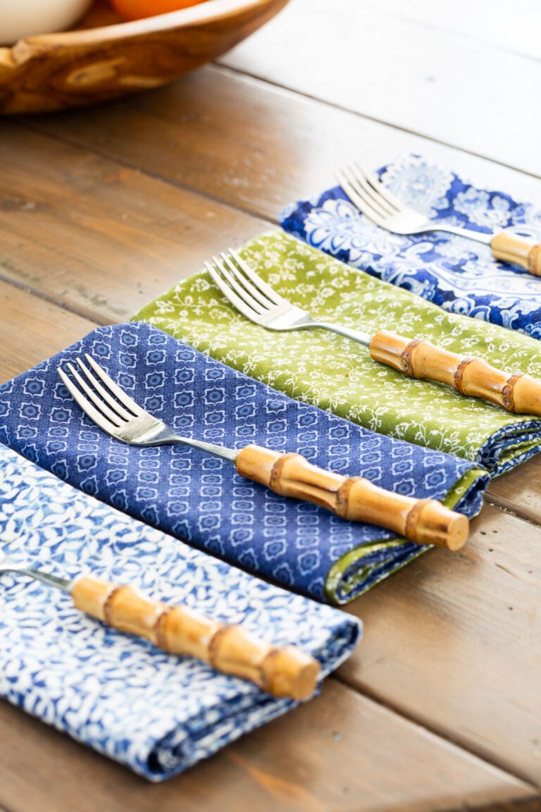 4 finished blue, green and white napkins folded and lined up on a table with a fork on each.